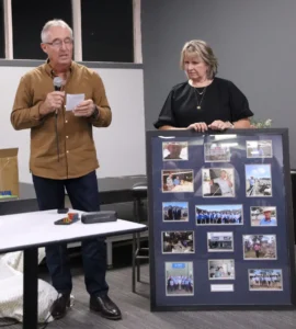 A man in a brown shirt (Damien Harrington) speaks into a microphone, holding notes, next to a woman in a black top who stands beside a large framed photo collage. This image captures Damien Harrington's farewell speech at an AWN Wimmera Livestock and Property event, celebrating his career.