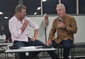 Wayne Driscoll holds a microphone, speaking to an audience, while Damien Harrington listens, sitting at a table. This image is from a farewell event for Damien Harrington, an AWN Wimmera livestock and property agent.