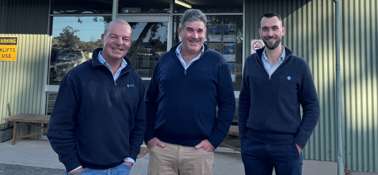 Three men standing and smiling in front of a corrugated iron building with AWN signage in the background. All are wearing collared shirts and dark pullovers with company logos.
