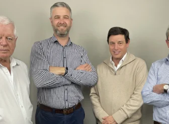 A professional studio portrait of four men (AWN Board members) standing side-by-side against a plain grey background. From left to right: Brendon Lunney (Deputy Chairman), a man with white hair wearing a white collared shirt; Rick Maybury (MD & CEO), a man with short grey hair and a beard wearing a blue and white checkered button-up shirt with his arms crossed; John Colley (Chairman), a man with dark hair wearing a light brown collared sweater; and Ben Barlow (Non-Executive Director), a man with glasses and grey hair wearing a blue and white striped collared shirt with his arms crossed.