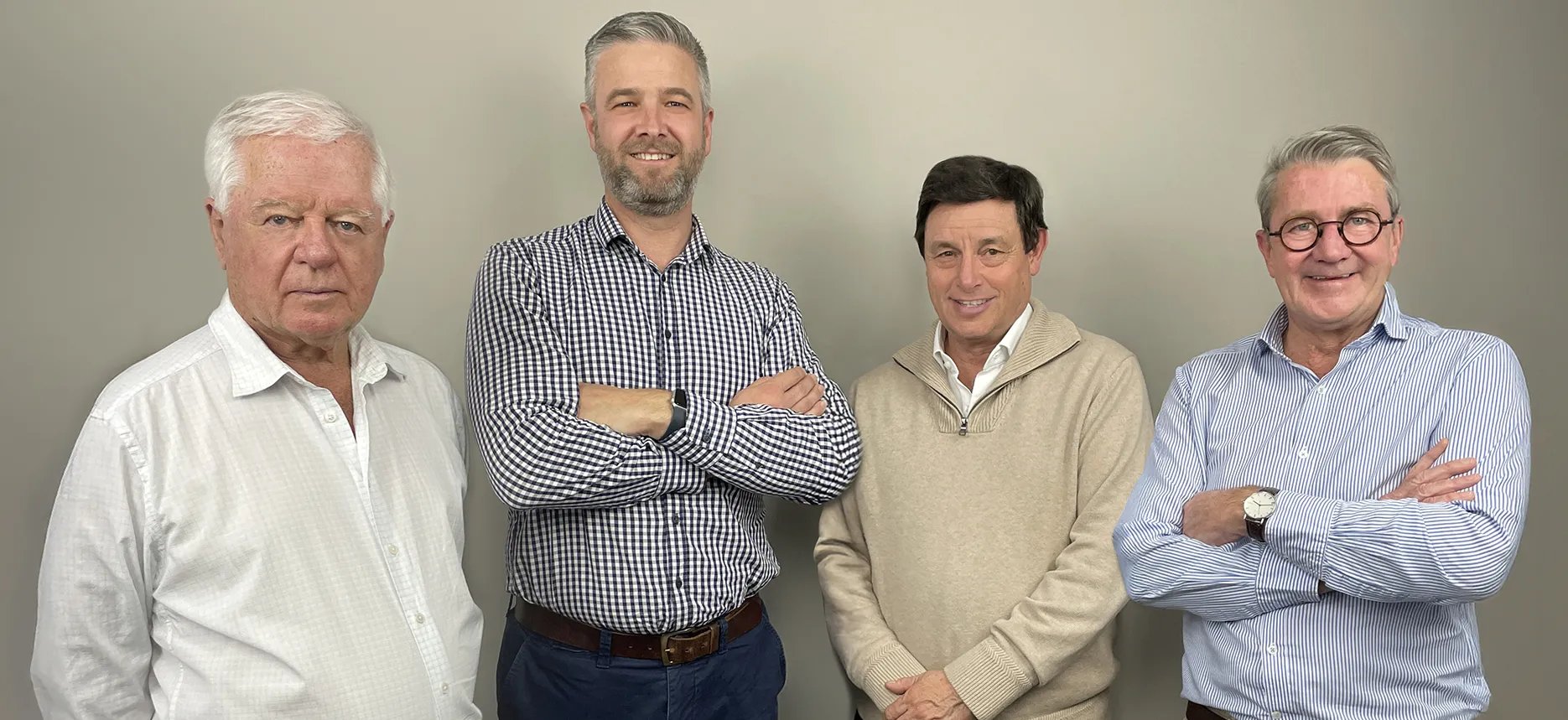 A professional studio portrait of four men (AWN Board members) standing side-by-side against a plain grey background. From left to right: Brendon Lunney (Deputy Chairman), a man with white hair wearing a white collared shirt; Rick Maybury (MD & CEO), a man with short grey hair and a beard wearing a blue and white checkered button-up shirt with his arms crossed; John Colley (Chairman), a man with dark hair wearing a light brown collared sweater; and Ben Barlow (Non-Executive Director), a man with glasses and grey hair wearing a blue and white striped collared shirt with his arms crossed.