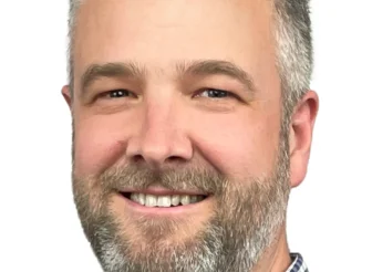 A professional headshot of Rick Maybury, MD & CEO of AWN, smiling confidently. He is a man with short, greying hair and a beard, wearing a blue and white checkered collared shirt, against a plain white background.