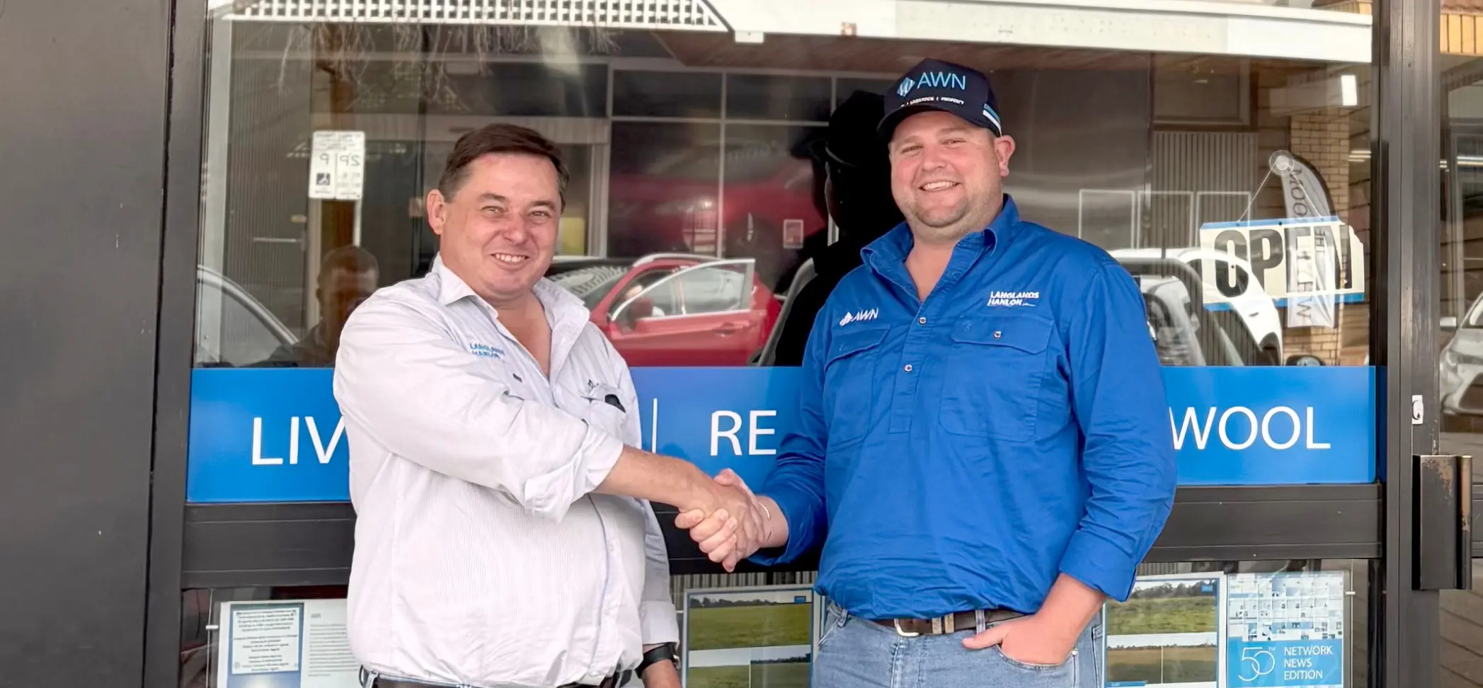 A horizontal, full-body shot of two men, Greg Miller and Cooper Byrnes, shaking hands in front of the AWN Langlands Hanlon branch office in Parkes, NSW. The storefront windows are covered with promotional posters. Both men are wearing collared shirts and jeans.