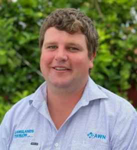 A headshot of Cooper Byrnes, the AWN Parkes and Langlands Hanlon branch manager. He is a young man with brown hair, smiling and wearing a blue and white striped work shirt with the AWN and Langlands Hanlon logos on it. The background is a green hedge.