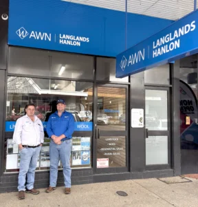 A vertical, full-body shot of two men, Greg Miller and Cooper Byrnes, standing side-by-side in front of the AWN Langlands Hanlon branch office in Parkes, NSW. The storefront has a large blue awning with the AWN and Langlands Hanlon logos. Both men are wearing collared shirts and jeans.